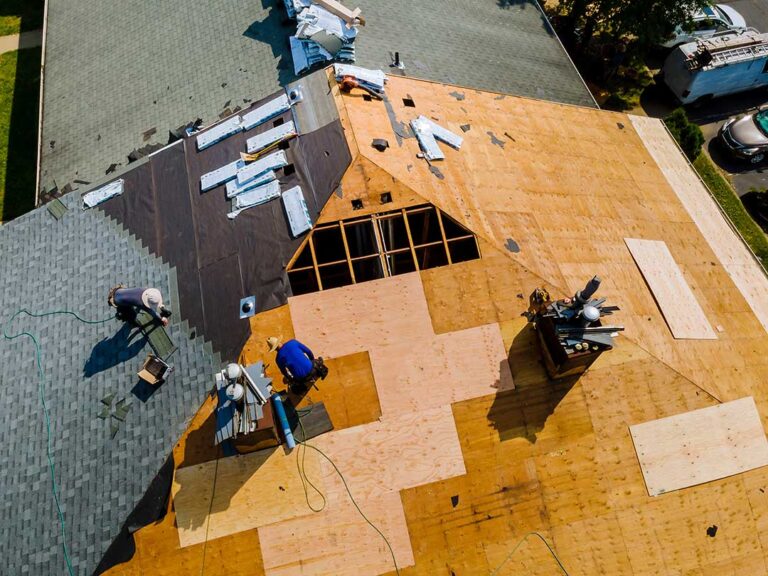 A worker installing a roof cricket behind a brick chimney on an asphalt shingle roof