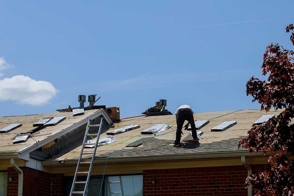 A roofer installs metal flashing around a plumbing vent on a shingled roof.