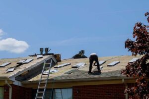 A roofer installs metal flashing around a plumbing vent on a shingled roof.